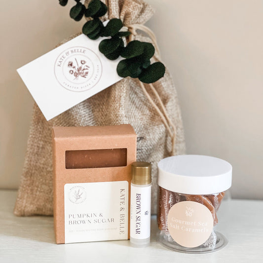 A curated gift set featuring a bar of oatmeal milk and honey soap in a kraft box, a small roller bottle, a clear jar of dried botanicals with a white lid, and a burlap drawstring bag with a white card and eucalyptus sprig, all on a light surface against a neutral beige background.